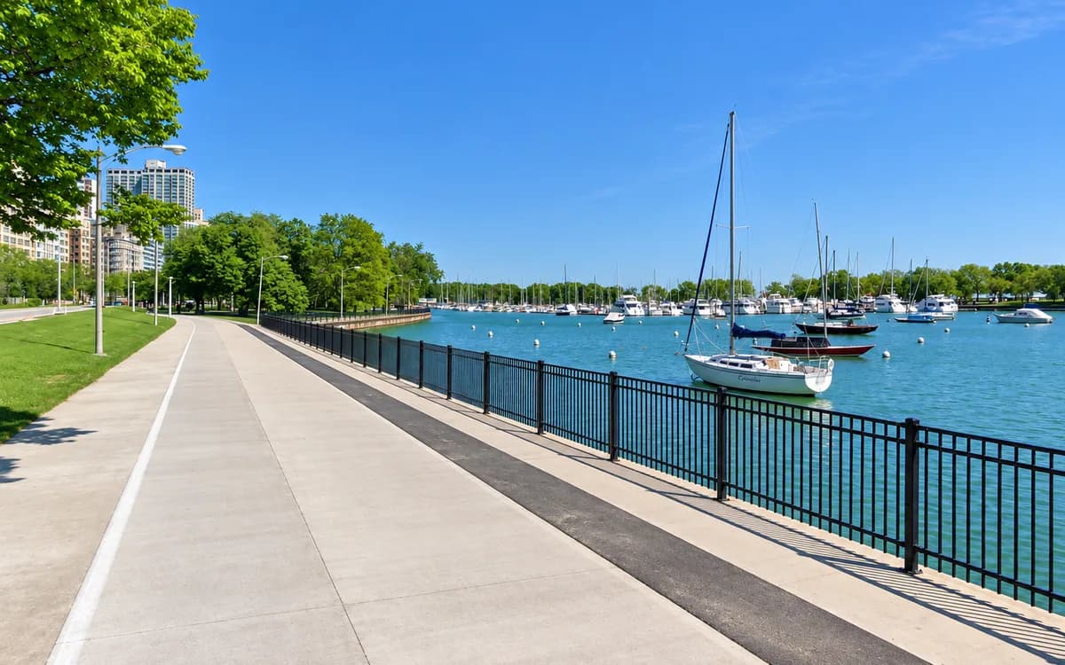 A paved lakefront trail beside Belmont Harbor, with sailboats moored on the water and the Chicago skyline in the distance